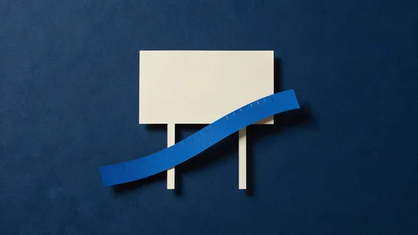 Un espectacular de papel crema sobre dos patas y una cinta métrica de papel azul cruzándolo en diagonal, sobre una superficie azul marino